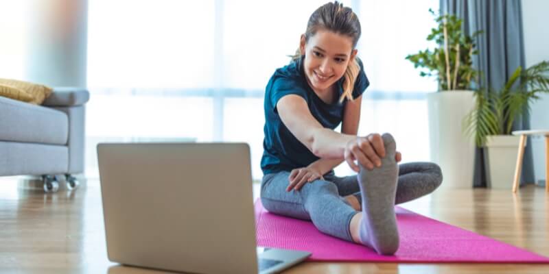 Young woman stretching on a yoga mat with a laptop for a class.